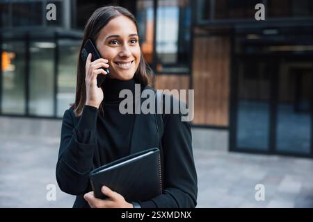 Lächelnde Frau, die schwarze formelle Kleidung mit hohem Kragen trägt. Geschäftsfrau, die auf ihrem Handy spricht, während sie wegblickt. Stockfoto