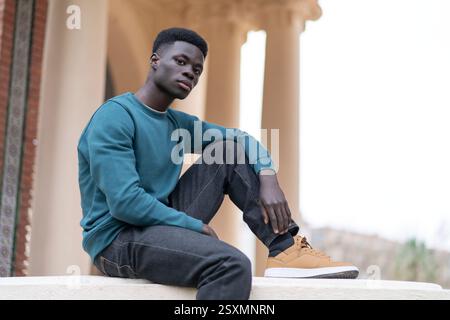Stilvoller junger Mann, der in einer urbanen Umgebung sitzt und moderne Mode und eine entspannte Pose präsentiert Stockfoto