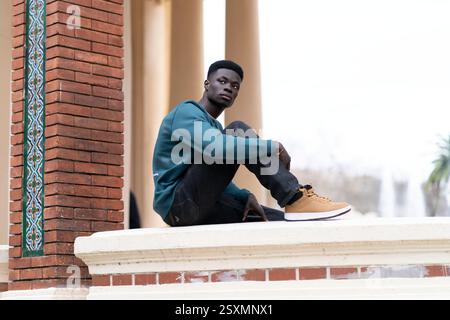 Nachdenklicher afroamerikanischer Mann, der auf einer niedrigen Wand sitzt, lässige Kleidung trägt und wegschaut Stockfoto