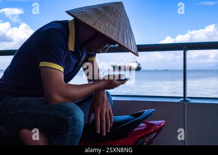 Ein Mann mit einem konischen Hut sitzt auf seinem Roller auf der Cat Hai - Cat Ba Fähre und schaut auf sein Handy, ein Frachtschiff, das in der Ferne verschwimmt. Stockfoto