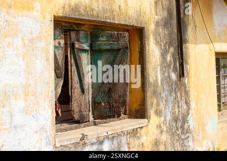 Ein altes, verwittertes Holzfenster mit abblätternder grüner Farbe in einer gelben, gerissenen Wand, die Verfall und rustikalen Charme zeigt. Stockfoto