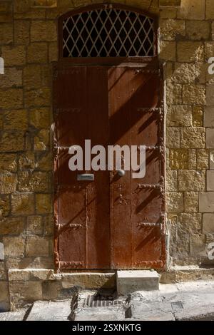 Stark seitlich beleuchteter hölzerner Eingang, mit einem Heckfenster, zu einem aus Kalkstein gebauten Grundstück auf der Insel Malta. Stockfoto