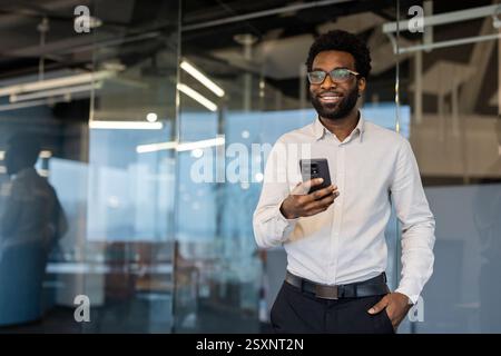 Erfolgreicher Geschäftsmann mit Telefon in der Hand, der im Büro am Arbeitsplatz steht. Mann, der zufrieden lächelt und aus dem Fenster blickt, Investor, der die App auf dem Smartphone nutzt. Stockfoto