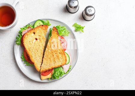 Club Sandwich halbiert und Tasse Tee auf weißem Hintergrund, Draufsicht, Kopierraum. Sandwich mit Schinken und Cheddar mit Tomaten, Gurke und Salat Stockfoto
