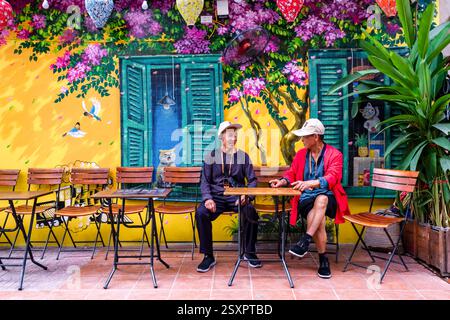 Zwei Männer, die Baseballmützen tragen, sitzen in einem Open-Air-Restaurant in der Train Street, die Wand dahinter ist mit farbenfrohen Graffiti verziert. Hanoi Ha Stockfoto