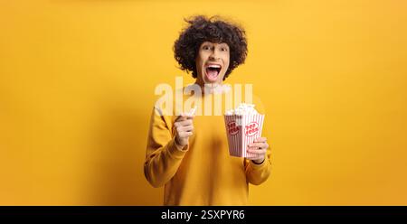 Überglückter junger Mann in gelbem Sweatshirt, der eine Kiste Popcorn auf gelbem Hintergrund hält Stockfoto