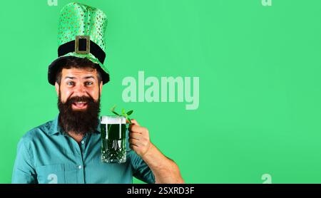Patrick Day Pub Party. Glücklicher bärtiger Mann in grünem Hemd und Hut, der grünes Bier trinkt. Irische Tradition. Ein bärtiger Mann mit Kobold-Hut hält einen Becher Stockfoto