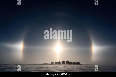 Sonnenhunde und ein Halo schaffen stimmungsvolle Optik über einer verschneiten Landschaft mit kleinen Häusern Stockfoto