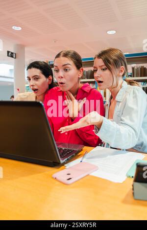 Drei überraschte Mädchen verbrachten einen aufregenden Moment, während sie zusammen in einer Bibliothek einen Laptop benutzten. Ihre Ausdrücke waren voller Staunen und Neugier, als sie etwas faszinierendes auf dem Bildschirm entdecken. Hochwertige Fotos Stockfoto