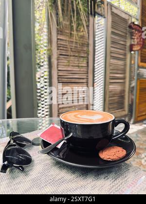 Eine gemütliche Café-Szene mit einer Tasse Kaffee mit Latte Art, einem Keks an der Seite und einer Sonnenbrille auf einem Tisch. Der Hintergrund besteht aus Holzteppich Stockfoto