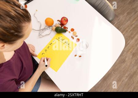 Eine Person führt sorgfältig Artikel für die Zubereitung von Mahlzeiten auf, umgeben von einer Vielzahl von frischem, farbenfrohen Obst und Gemüse Stockfoto
