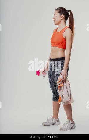 Junge sportliche Frau, die im Profil steht, eine Wasserflasche und ein Handtuch hält und nach einem belebenden Workout erfrischt und Energie ausstrahlt Stockfoto