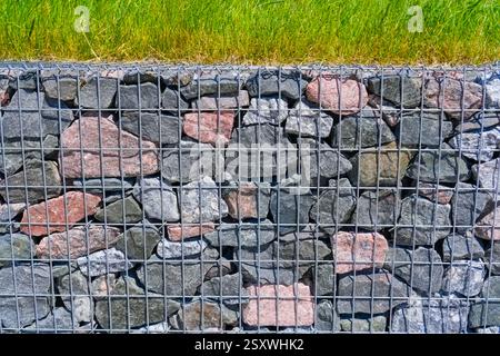 Eine Gabionenwand mit Metallgitterkäfigen, die mit bunten Steinen gefüllt sind, bildet eine stabile Barriere. Das Oberteil ist mit leuchtendem grünem Gras, Profi, verziert Stockfoto