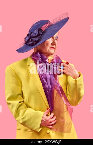 Ältere Frau in gelbem Blazer mit breiter Krempe und blauem Hut, der den purpurpurnen Schal anpasst, während sie vor rosa Hintergrund posiert. Stockfoto