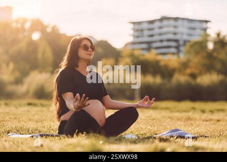 Junge schöne schwangere Frau mit Sonnenbrille macht Yoga auf der Matte draußen auf dem Feld. Stockfoto