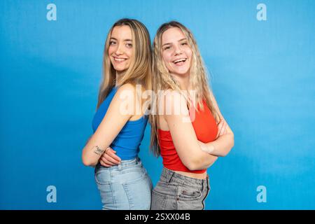 Zwei glückliche junge Frauen in lässiger Kleidung, die mit überkreuzten Armen hinter sich stehen und auf blauem Hintergrund lächeln Stockfoto