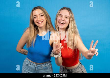 Zwei glückliche junge Frauen, die mit Fingern Friedenszeichen machen, lächeln und Spaß auf blauem Studiohintergrund haben Stockfoto