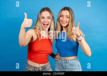 Zwei glückliche junge Frauen, die Daumen hoch halten und auf blauem Hintergrund lächeln Stockfoto