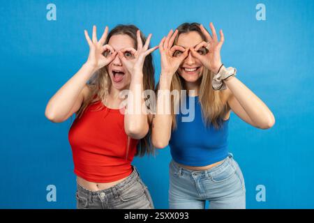 Zwei fröhliche Freundinnen machen Ferngläser mit ihren Fingern und haben Spaß vor einem leuchtend blauen Hintergrund Stockfoto