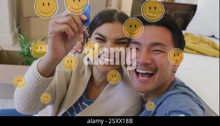 Bild von Smiley-Gesichter-Ikonen über glückliches birassisches Paar mit Hausschlüssel Stockfoto