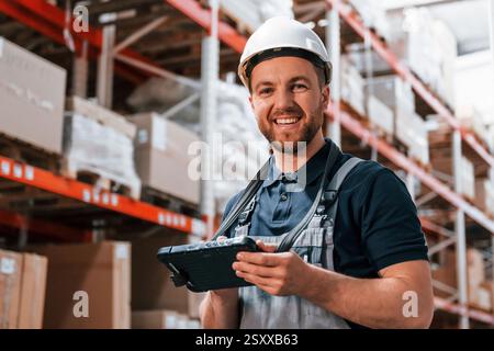 Mit weißem Schutzhelm und mit Tablette. Mitarbeiter in Uniform arbeitet tagsüber im Lager. Stockfoto