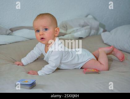 Ein 3-5 Monate alter Junge liegt auf seinem Bauch auf einem weißen Bett in einem blauen Body und schaut in die Kamera. Hochwertige Fotos Stockfoto