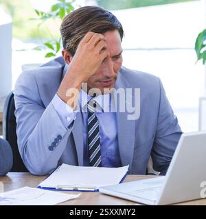 Im Büro, Geschäftsmann, der Dokumente analysiert und an einem Laptop arbeitet, sieht gestresst aus Stockfoto