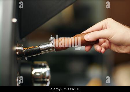 Mittlere Nahaufnahme einer nicht erkennbaren männlichen Hand, die einen kleinen Metalllöffel mit Holzgriff mit Kaffeebohnen hält und in die Maschine setzt Stockfoto