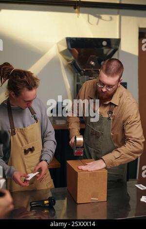Vertikal von zwei kaukasischen Arbeitern, die Pappkarton mit Klebeband verpacken und Etikett darauf anbringen Stockfoto