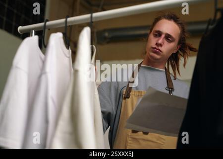 Niedrigwinkelaufnahme eines jungen erwachsenen Mannes mit Dreadlocks, der Papiere hält und übergroße T-Shirts an Kleiderbügeln ansieht Stockfoto