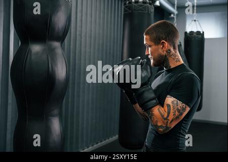 Mit Lochbeutel. Der junge tätowierte Mann übt im Fitnessstudio das Boxen. Stockfoto