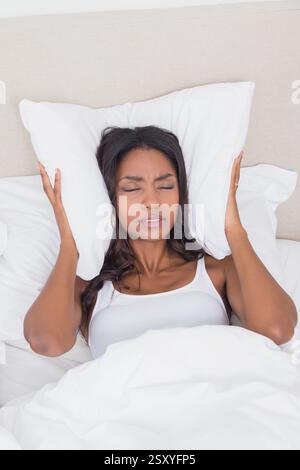 Afroamerikanerin im Bett, die Ohren mit Kissen bedeckt und frustriert aussieht Stockfoto