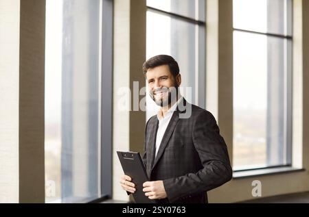 Junger, lächelnder, attraktiver Unternehmer im formellen Anzug, der im modernen Büro steht. Stockfoto