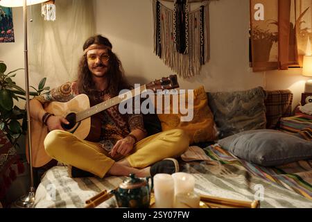 Lächelnder junger Mann, der Akustikgitarre in einem gemütlichen Zimmer spielt Stockfoto