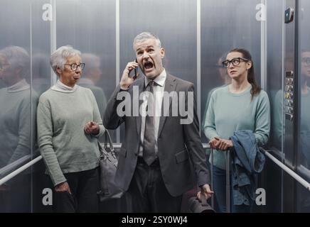 Ein gestresster wütender Geschäftsmann, der im Aufzug in sein Smartphone schreit, zwei Frauen sind genervt und schauen sich gegenseitig an Stockfoto