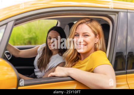 Eine lächelnde blonde Frau lehnt sich aus einem gelben Taxifenster, während ihr brünette Freund auf dem Fahrersitz sitzt Stockfoto