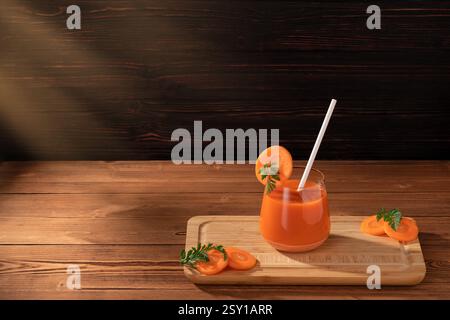 Holztisch unter warmen Sonnenstrahlen. Glasbecher mit Stroh und frischem Karottensaft auf Schneidebrett. Stockfoto