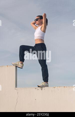 Eine sportliche Frau, die an einer Wand steht, ihre Haare bindet, Trainingshosen trägt Stockfoto
