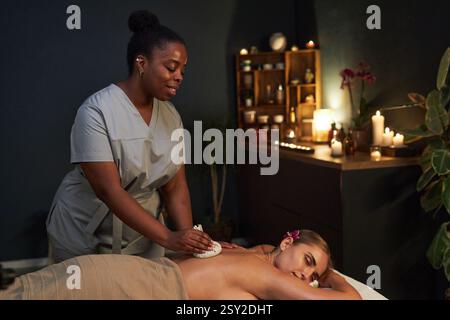 Massagetherapeut übt entspannende Massagetherapie auf Kunden, die sich in einem gemütlichen Raum mit Kerzen, Pflanzen und Holzregalen mit Gläsern und Behältern liegen Stockfoto