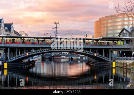 Panoramablick Berliner Bahnhof Friedrichstraße Zug überquert die Metallbrücke vor dem dramatischen warmen Sonnenuntergangshimmel. Abends Berlin Mitte Spree Szene Urban Stockfoto