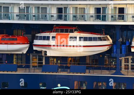 Detaillierte Sicht auf das Äußere eines Kreuzfahrtschiffes mit zwei Rettungsbooten, verglasten Balkonen mit Möbeln und einem tiefblauen Rumpf mit weiß-roten Akzenten Stockfoto