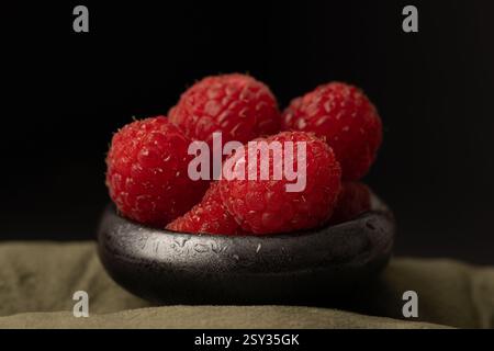 Frische Himbeeren auf einer winzigen, schwarz glänzenden Schale und auf einer grünen Tischdecke. Dramatische Studiobeleuchtung. Stockfoto