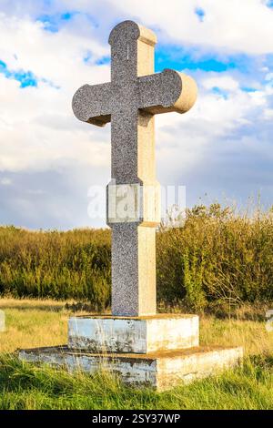 Großes Steinkreuz steht auf einem Feld. Das Kreuz ist von Gras und Bäumen umgeben. Der Himmel ist bewölkt und die Sonne scheint Stockfoto