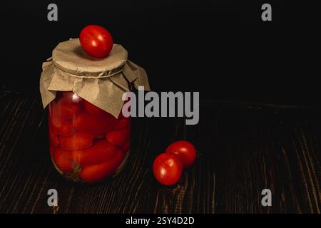 Ein Glas mit hausgemachten eingelegten Tomaten steht auf einer dunklen Holzoberfläche und zeigt leuchtend rote Kirschtomaten. Weitere Tomaten liegen verstreut Stockfoto