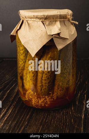 Ein Glasgefäß mit hausgemachten eingelegten Gurken steht auf einer Holzoberfläche. Das rustikale Glas mit braunem Papier zeigt die lebendigen Gurken, r Stockfoto
