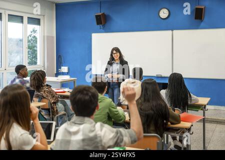 Multiethnische Schüler heben während eines Unterrichts von Lehrerinnen in der Oberstufe die Hände Stockfoto
