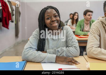 Die junge schwarze Schülerin lächelt, während sie an ihrem Schreibtisch in einem Klassenzimmer sitzt, während sie mit ihren Klassenkameraden unterrichtet Stockfoto