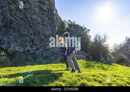 Zwei weibliche Klettererinnen laufen auf einem grasbewachsenen Hügel in der Nähe einer großen Felswände, tragen Kletterausrüstung und tragen Helme Stockfoto