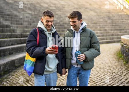 Glückliches schwules Paar, das Hand in Hand geht, lächelt und ihr Smartphone überprüft, während es einen Wintertag im Freien in der Stadt genießt Stockfoto