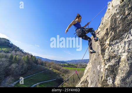 Klettererin, die eine Felswände hinaufsteigt, einen sonnigen Tag und die wunderschöne Aussicht auf das Tal darunter genießt Stockfoto
